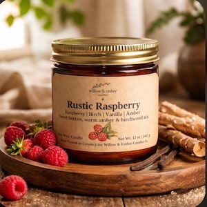 May include: A glass jar candle with a gold lid, labeled "Rustic Raspberry." The label lists ingredients and scent notes. Fresh raspberries and vanilla beans are arranged around the candle on a wooden tray. The candle weighs 12 oz.