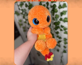 May include: A crocheted fire salamander plush toy with orange and yellow fur, blue eyes, and a red tail. The toy is being held by a hand.