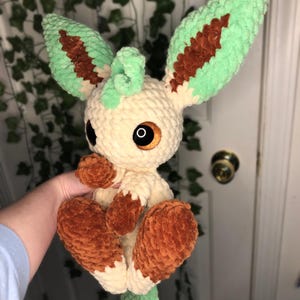 Op de afbeelding: Een gehaakt pluche speelgoed van een lichtbruine Eevee met groene en bruine oren en een groene staart. Het speelgoed heeft grote bruine ogen en een klein, gekruld groen hoorn op zijn hoofd.