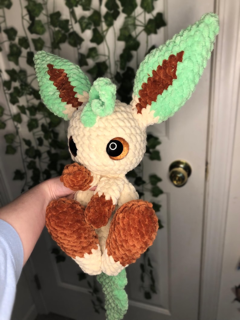 Op de afbeelding: Een gehaakt pluche speelgoed van een lichtbruine Eevee met groene en bruine oren en een groene staart. Het speelgoed heeft grote bruine ogen en een klein, gekruld groen hoorn op zijn hoofd.