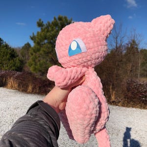 May include: A pink crocheted plush toy of a cartoon character with a large head, small ears, and a long tail. The toy has blue and white eye details. The plush is held up against a background of trees and a blue sky.