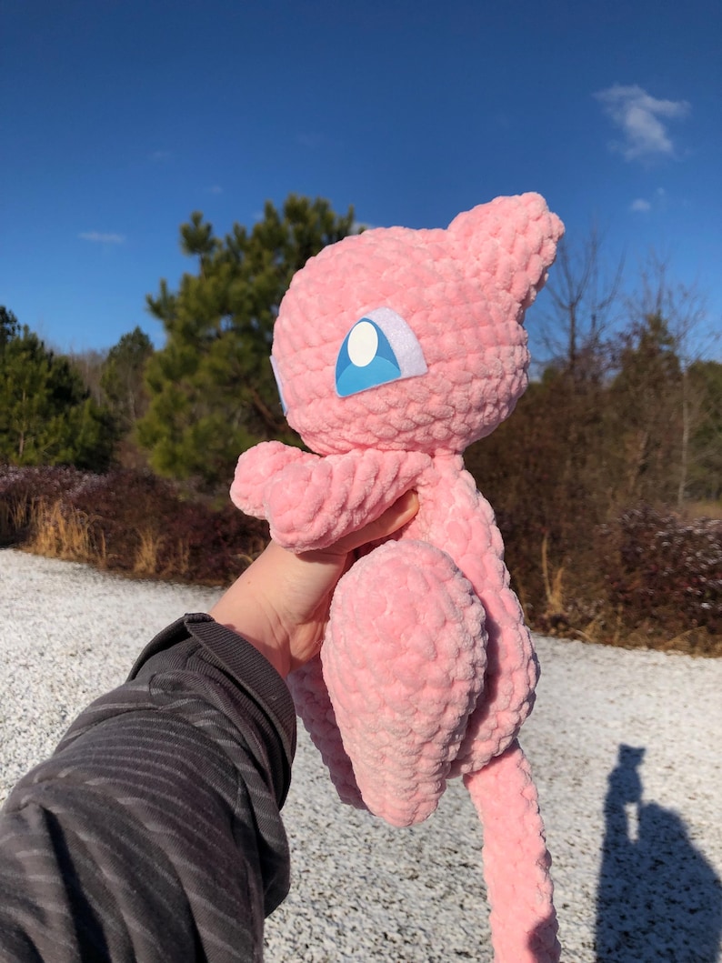 May include: A pink crocheted plush toy of a cartoon character with a large head, small ears, and a long tail. The toy has blue and white eye details. The plush is held up against a background of trees and a blue sky.