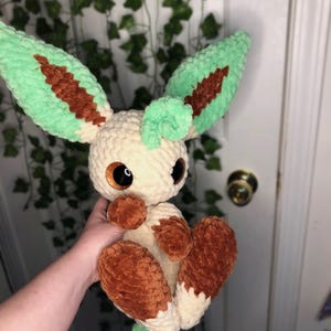 Op de afbeelding: Een gehaakt pluche speelgoed dat lijkt op een Pokemon-personage. Het speelgoed is cr&egrave;mekleurig met bruine accenten en groene oren en staart. Het speelgoed heeft grote, ronde, bruine ogen.