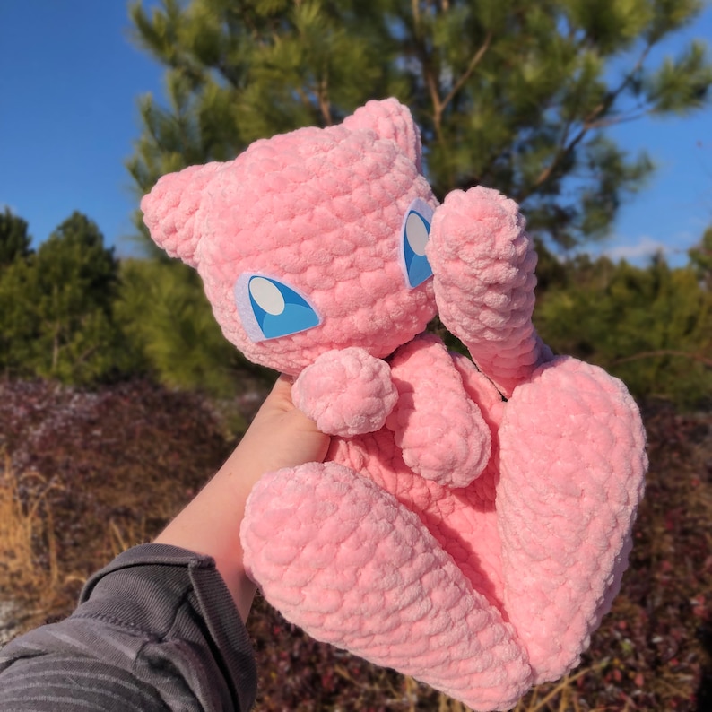 May include: A pink, plush, crocheted Mew doll with blue eyes. The doll has large feet and ears, and is held up against a background of green trees and a blue sky. The doll is made of a soft, textured yarn.