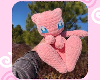 May include: A pink, crocheted Mew plushie with blue eyes and a small nose. The plushie is made of a soft, textured yarn and has a cute, friendly expression. The image includes the text "PDF Pattern" and "Mew".