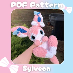 Pode incluir: Um padrão de crochê para um brinquedo de pelúcia Sylveon Pokémon. O padrão é para um Sylveon rosa e branco com detalhes azuis e azul claro. O padrão está disponível para download em PDF.