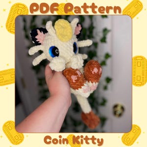 May include: A handmade crochet Coin Kitty plushie in cream, brown, and pink. The plushie has blue eyes, black and pink ears, and a yellow head crest. The image includes the text "PDF Pattern" and "Coin Kitty".
