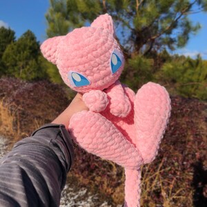 May include: A pink crocheted plush toy with large blue eyes and a long tail. The toy is held up against a backdrop of trees and a blue sky. The plush has a soft, textured appearance.