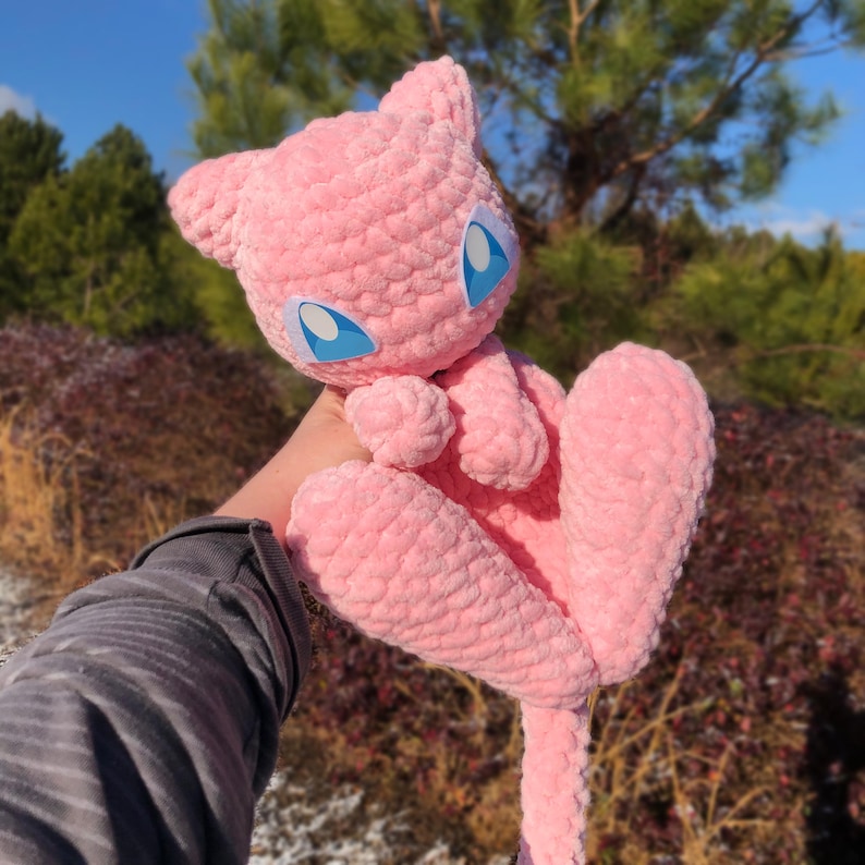 May include: A pink crocheted plush toy with large blue eyes and a long tail. The toy is held up against a backdrop of trees and a blue sky. The plush has a soft, textured appearance.