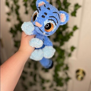 May include: A handmade blue plush tiger cub with light blue accents. The tiger cub has large brown eyes, pink nose and ears, and black stripes. The plush toy is made of soft, textured material.
