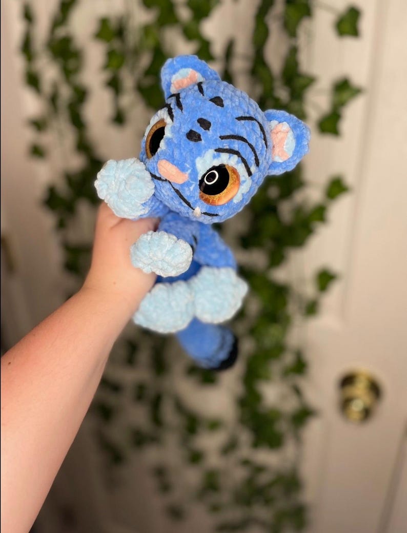 May include: A handmade blue plush tiger cub with light blue accents. The tiger cub has large brown eyes, pink nose and ears, and black stripes. The plush toy is made of soft, textured material.