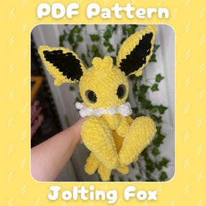 May include: A crocheted yellow creature with black-tipped ears and large eyes. It has a white collar and large feet. The text "PDF Pattern" and "Jolting Fox" are displayed on a yellow background. The plush toy is handmade.