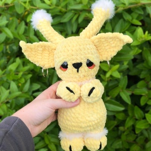 May include: A crocheted plush toy in the shape of a yellow creature with large ears, white fur accents, and black eyes. The toy is holding its hands together in front of its chest.