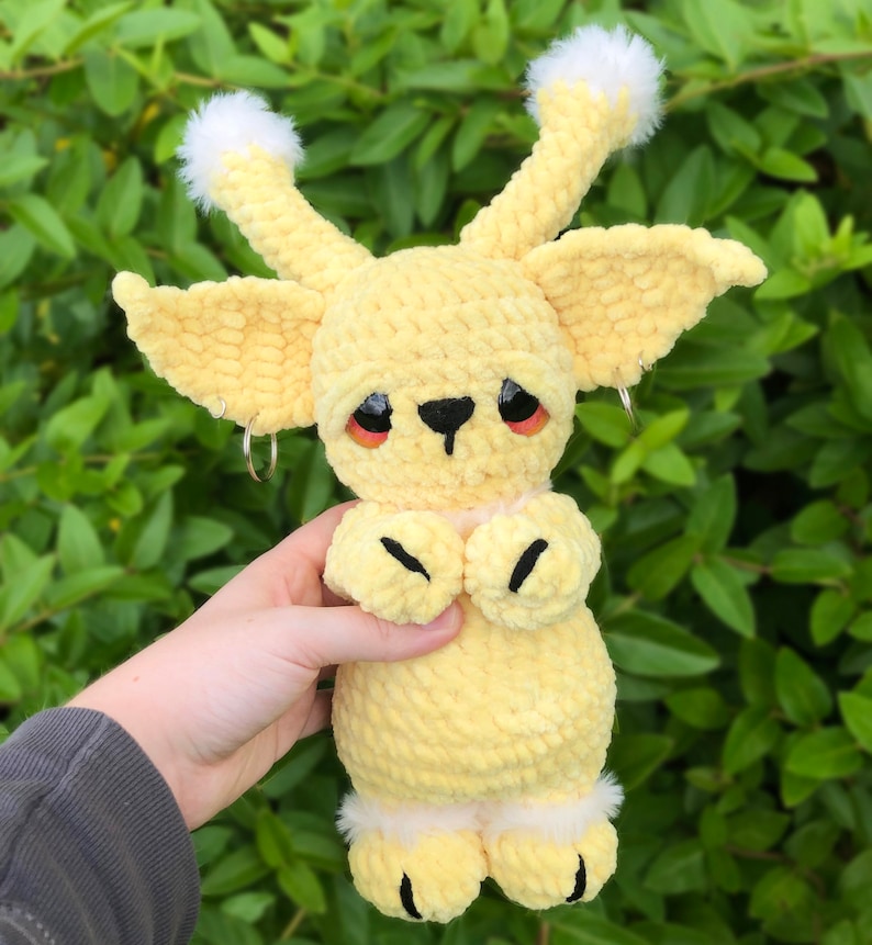 May include: A crocheted plush toy in the shape of a yellow creature with large ears, white fur accents, and black eyes. The toy is holding its hands together in front of its chest.