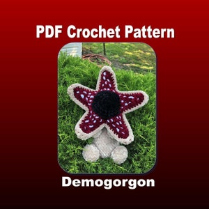 May include: A crochet pattern for a Demogorgon from the show Stranger Things. The Demogorgon is a red and white crocheted star with a black center. The Demogorgon is sitting on a bed of green grass.