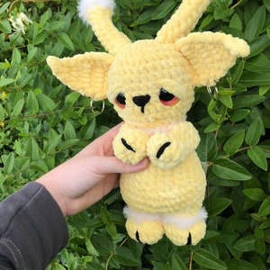 May include: A yellow crocheted plush toy with white fur accents and black eyes. The toy has a small black nose and black crocheted paws.