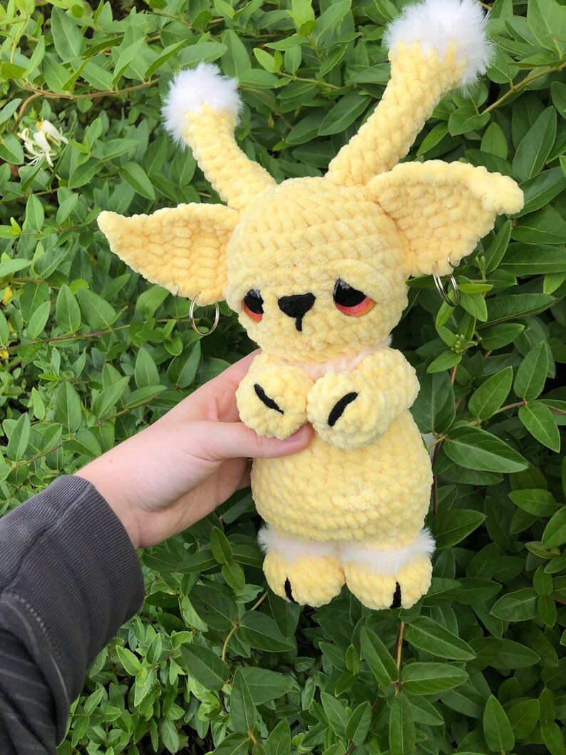 May include: A yellow crocheted plush toy with white fur accents and black eyes. The toy has a small black nose and black crocheted paws.