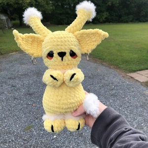 May include: A crocheted yellow plush toy with white fur accents. The toy has large ears, a small black nose, and black eyes. The toy is holding its paws together in front of its chest.