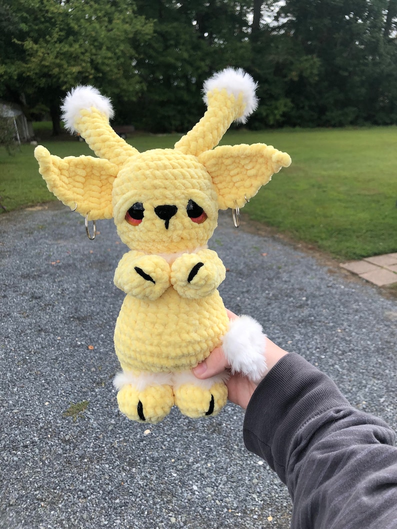 May include: A crocheted yellow plush toy with white fur accents. The toy has large ears, a small black nose, and black eyes. The toy is holding its paws together in front of its chest.