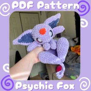 May include: A crocheted plush toy in the shape of a purple and blue psychic fox. The toy has large white eyes and an orange nose. The toy is being held in a person's hand.