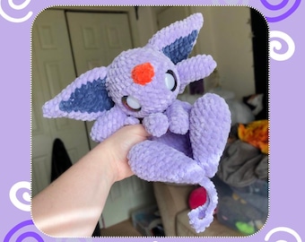 May include: A crocheted plush toy in the shape of a purple and blue psychic fox. The toy has large white eyes and an orange nose. The toy is being held in a person's hand.
