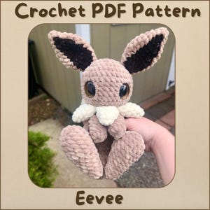 May include: Crochet PDF pattern for an Eevee plush toy. The Eevee is brown with brown ears and white fur around its neck. The Eevee is sitting in a person's hand.