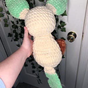 Op de afbeelding: Een gehaakt pluche speelgoed in de vorm van een lichtbruine Eevee met groene oren en staart. Het speelgoed wordt vastgehouden door een hand.