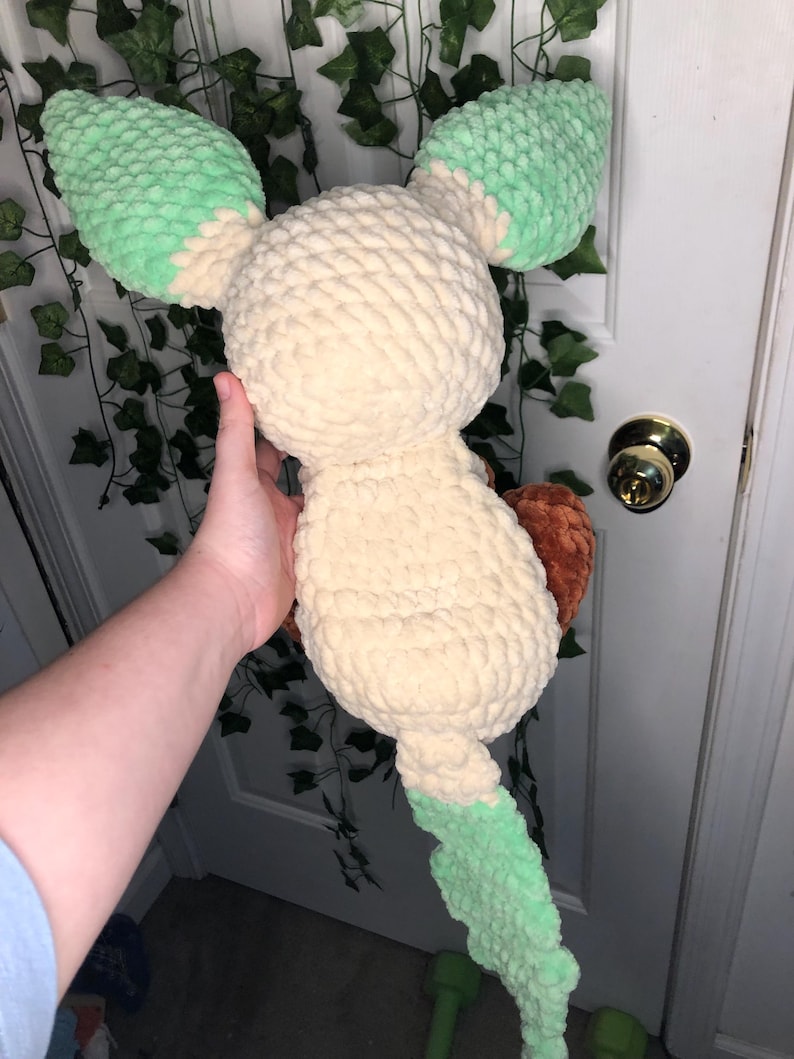 Op de afbeelding: Een gehaakt pluche speelgoed in de vorm van een lichtbruine Eevee met groene oren en staart. Het speelgoed wordt vastgehouden door een hand.