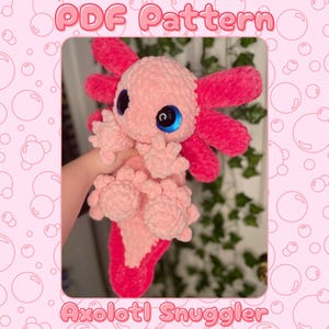 Könnte beinhalten: Ein rosa Axolotl-Plüschtier mit leuchtend rosa Kiemen und Akzenten sowie großen blauen Augen. Der Text "PDF Pattern" steht oben und "Axolotl Snuggler" unten. Der Hintergrund ist rosa mit Blasendesigns.