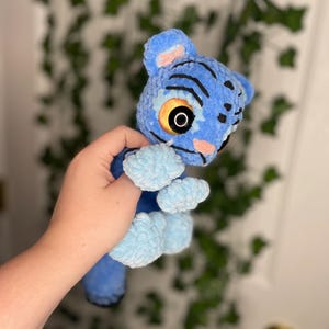 May include: A blue plush toy resembling a creature from the movie Avatar. The toy has black stripes, large yellow eyes, and light blue paws. The plush is made of a soft, textured material.