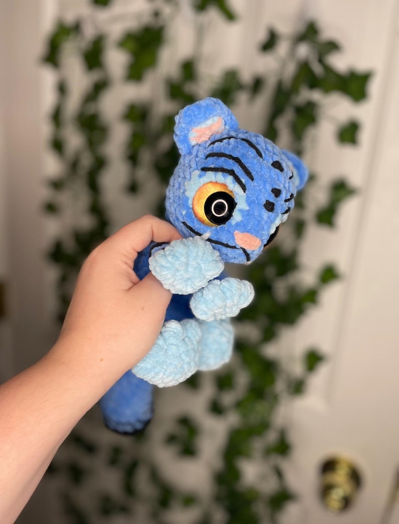 May include: A blue plush toy resembling a creature from the movie Avatar. The toy has black stripes, large yellow eyes, and light blue paws. The plush is made of a soft, textured material.