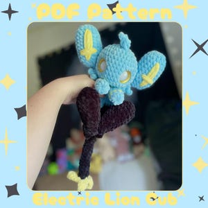 Puede incluir: Un peluche hecho a mano de ganchillo azul claro de Electric Lion Cub con detalles amarillos. El peluche tiene orejas grandes, una cola pequeña y patas marrones oscuras. El texto "PDF Pattern" y "Electric Lion Cub" son visibles.