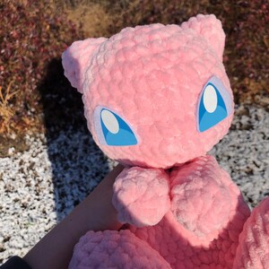 May include: A pink plush toy, resembling a cartoon character. The toy has large blue eyes with white highlights and small ears. The plush is made of a soft, textured material.