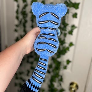 May include: A blue plush toy with black and white accents. The toy has a rounded head with two small ears, a segmented body, and a long tail. The tail is black at the end. The toy is held in a person's hand.