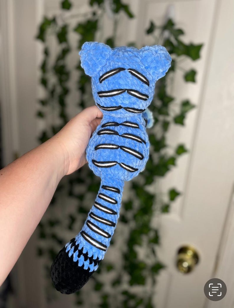 May include: A blue plush toy with black and white accents. The toy has a rounded head with two small ears, a segmented body, and a long tail. The tail is black at the end. The toy is held in a person's hand.
