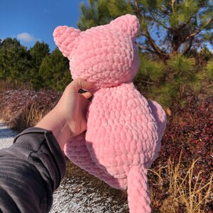 May include: A handmade pink crochet cat plush toy with a textured, soft appearance. The cat has pointed ears and a long tail. The plush toy is held up against a backdrop of trees and a blue sky.