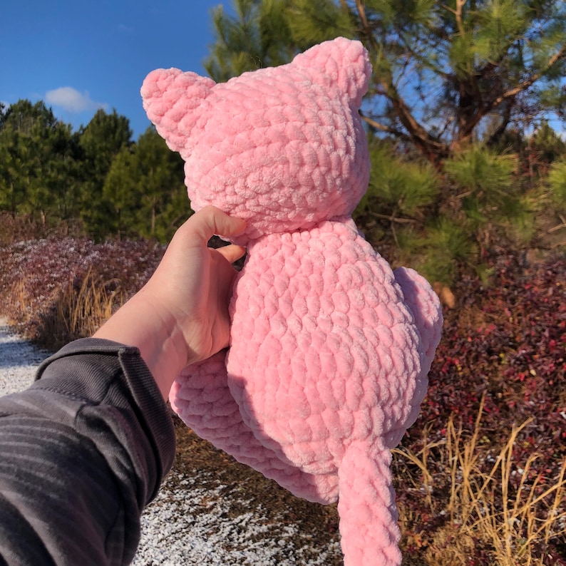 May include: A handmade pink crochet cat plush toy with a textured, soft appearance. The cat has pointed ears and a long tail. The plush toy is held up against a backdrop of trees and a blue sky.