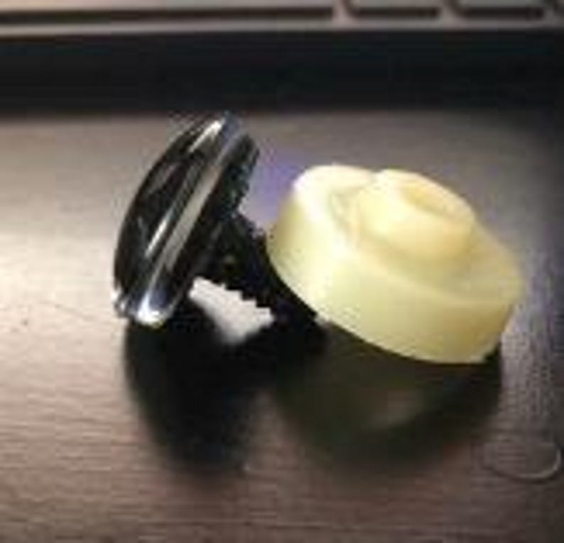 Op de afbeelding: Een zwarte plastic schroef met een zilveren ring en een witte plastic ring. De schroef heeft een platte kop en een kruiskop.