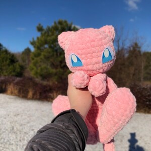 May include: A pink, plush Mew doll with blue eyes and a white outline is held up against a blue sky. The doll has a soft, textured appearance and is made of a fuzzy material. The background includes trees and a snowy ground.