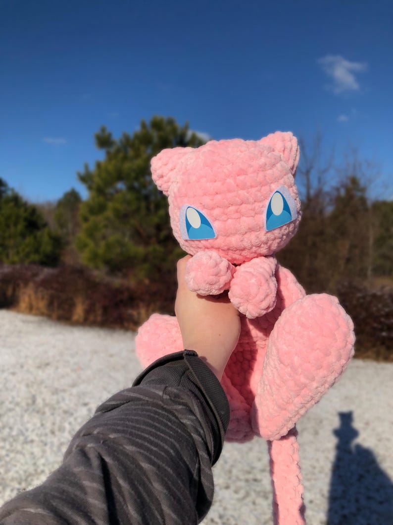 May include: A pink, plush Mew doll with blue eyes and a white outline is held up against a blue sky. The doll has a soft, textured appearance and is made of a fuzzy material. The background includes trees and a snowy ground.