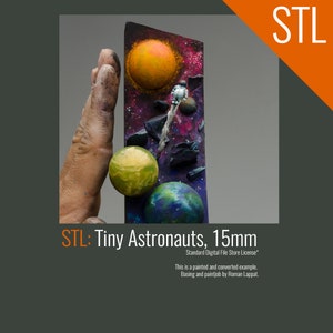 May include: A hand holds a small, painted model of a space scene with a tiny astronaut, planets, and a star. The model is 15mm tall and is made of a dark material. The background is a dark gray.