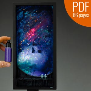 May include: A framed artwork depicting a galaxy with a swirling blue, green, and pink nebula. The galaxy is filled with white glitter and two small astronaut figurines are standing on a platform in the foreground. The artwork is in a black frame and is being held by a hand holding a bottle of purple paint.
