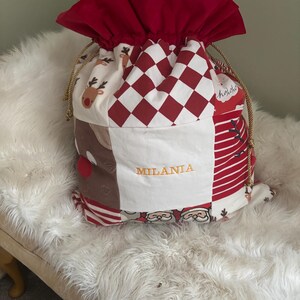 May include: A festive Christmas gift bag with a red drawstring top. The patchwork design includes reindeer, Santa Claus, and a red and white chequered pattern. The name "MILANIA" is embroidered in gold thread on the front of the bag.