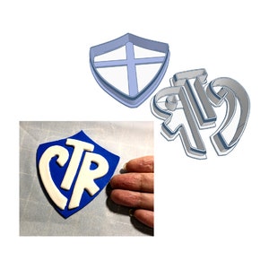 May include: A blue and white cookie cutter in the shape of a shield with the letters CTR inside. The cookie cutter is next to a blue and white cookie with the same design.