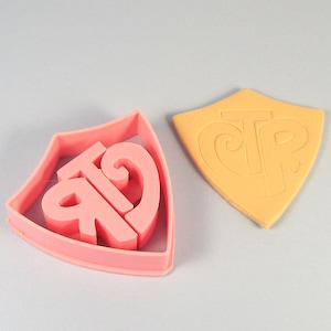 May include: Pink plastic cookie cutter shaped like a shield with the letters "CTR" inside. A separate orange cookie cutter with the same shape and letters is shown next to it.