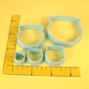 May include: Set of five blue plastic cookie cutters shaped like cauldrons, with the largest measuring 2 inches in diameter, and the smallest measuring 1 inch in diameter. The cutters are arranged on a yellow surface with a ruler for scale.
