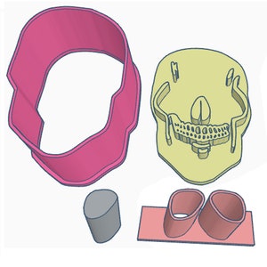May include: A set of four cookie cutters in the shape of a skull, two small circles, and a larger circle. The skull cutter is yellow, the circles are pink and gray, and the larger circle is pink.