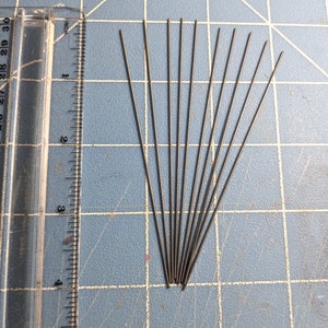May include: A close-up of ten thin, silver-colored metal rods arranged in a fan shape on a blue grid surface. A ruler is visible on the left side of the image.