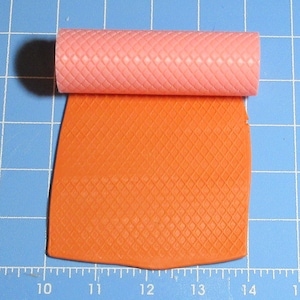 May include: A pink rolling pin with a diamond pattern is rolling over a sheet of orange fondant with a diamond pattern.