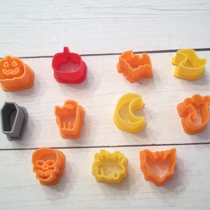 May include: A set of 12 Halloween-themed cookie cutters. The cutters are made of plastic and come in a variety of colors, including orange, yellow, gray, and red. The cutters feature designs such as a pumpkin, a bat, a ghost, a witch's hat, a coffin, a skull, a spider, a crescent moon, a cat, and a spider web.